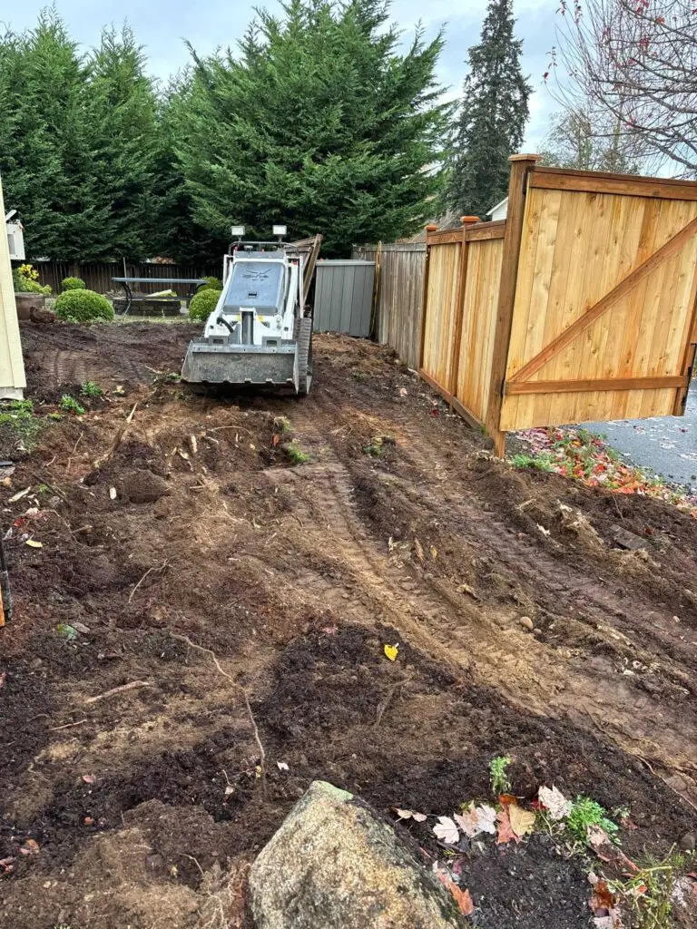 Residential excavation with a mini skid steer clearing and grading soil next to a new wooden fence.