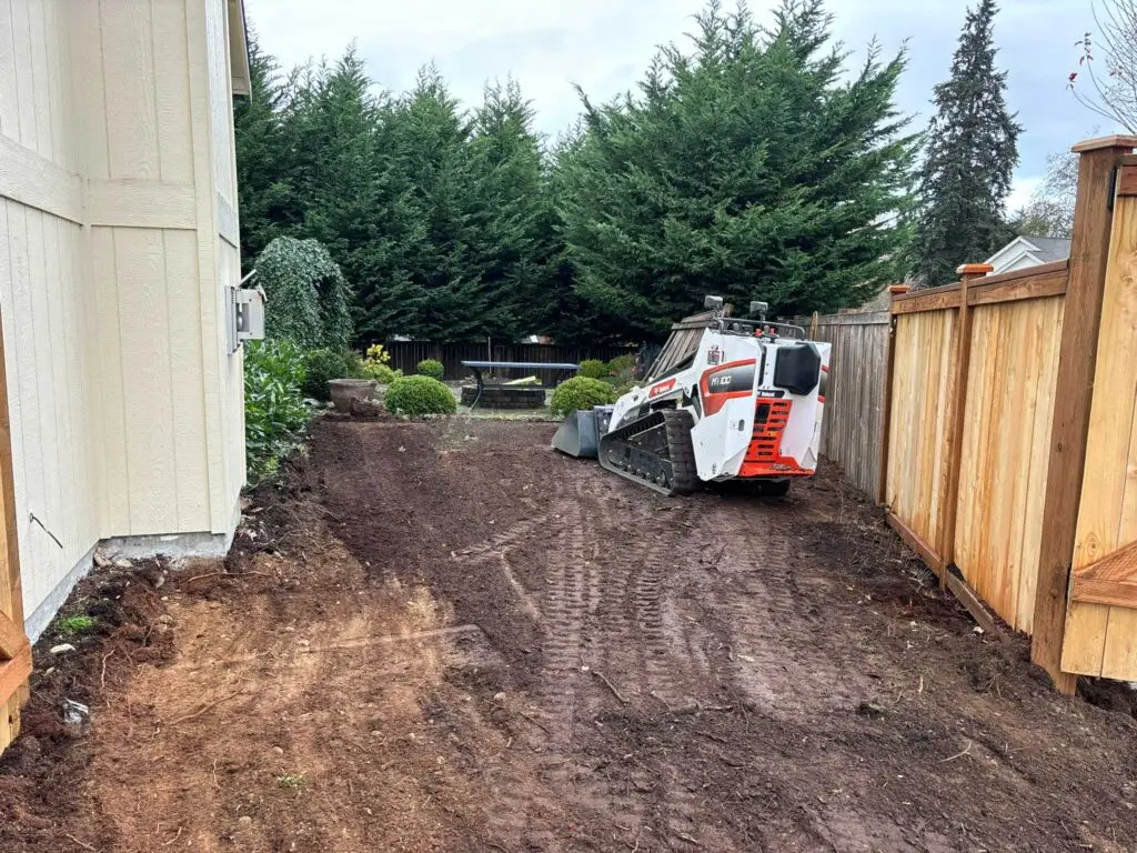 Mini skid steer grading and leveling soil along a residential home and wooden fence.