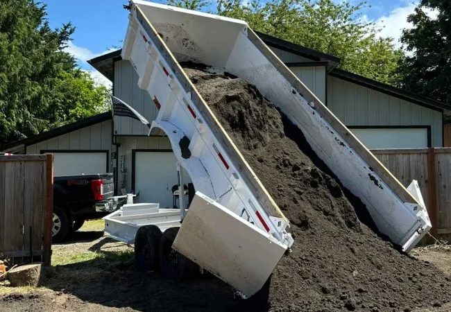 Dump trailer unloading a large load of topsoil in a residential driveway.